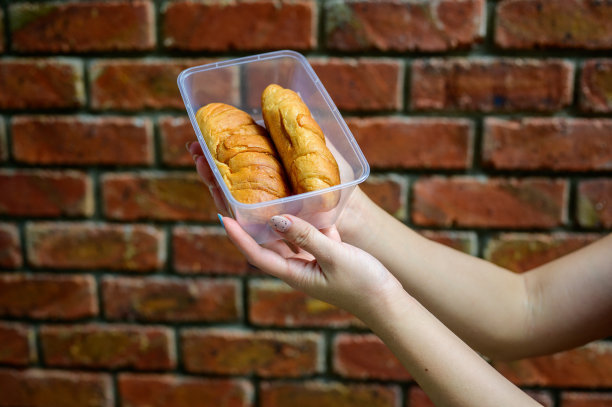 女人的手拿着一个塑料盒子与圆面包和牛角面包与砖墙背景图片下载
