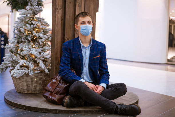 一名男子戴着面具坐在购物中心的圣诞树旁，手里拿着手机图片下载