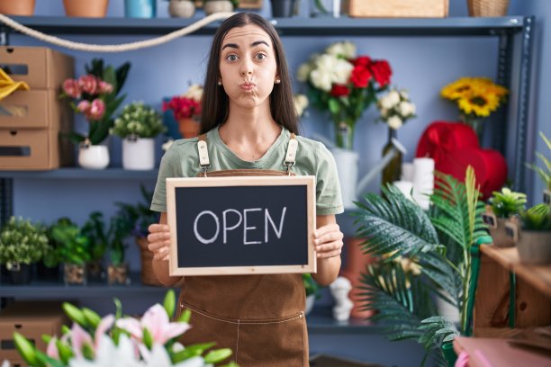 年轻的深色头发的女人在花店工作，拿着打开的标志，吹着滑稽的脸。嘴充满了空气，呼吸空气。图片下载