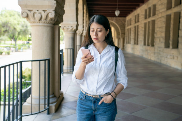 在美国加州一所学校大学的罗马式走廊上，一名严肃的亚裔台湾女大学生手插在口袋里，用手机阅读课程信息图片下载
