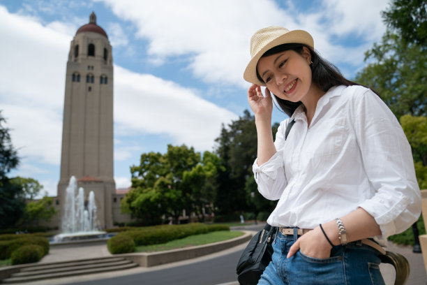 亚裔韩国女性游客的肖像带着害羞的微笑，在胡佛喷泉和钟楼的背景下摆姿势，手放在帽子和口袋在学校校园图片下载