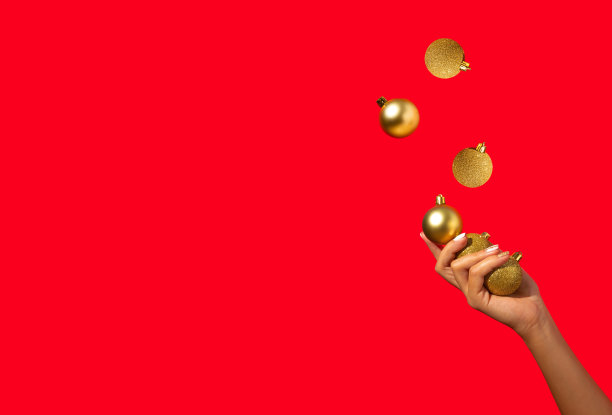 女人的手杂耍在红色背景上的金色圣诞装饰物。最小的假期概念。新年快乐。副本的空间。图片下载