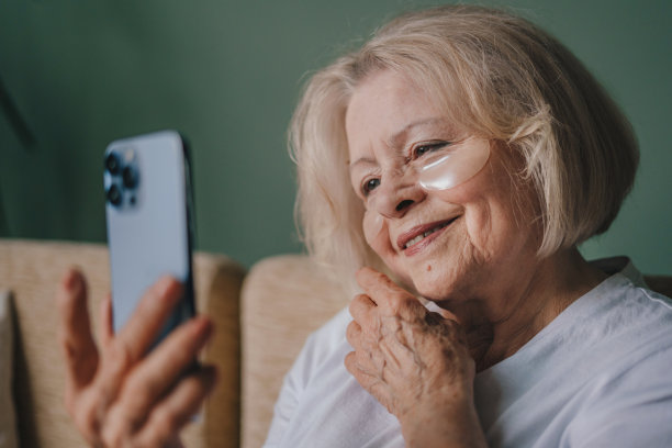 美丽的老年妇女为浮肿戴眼罩，用手机屏幕而不是镜子看她的样子。美容治疗。快乐的生活方式。美丽的肖像。自然美景。皮肤护理。图片下载