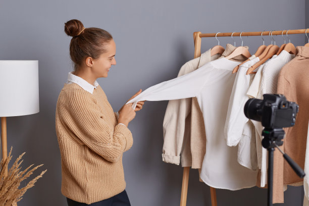 侧视图肖像黑头发年轻成年妇女与发髻记录视频相机与三脚架，显示衣服挂在架子上，谈论基本衣柜。图片下载
