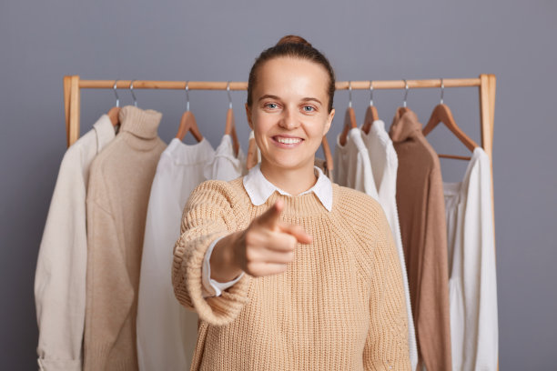 室内拍摄，迷人的女时尚顾问站在挂在架子上的衣服旁边，手指对着相机，邀请你去她的精品店或陈列室购物。图片下载