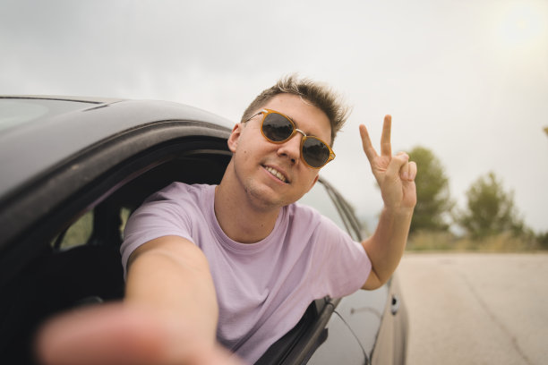 快乐的年轻司机在车里自拍度假-快乐的人在度假时租了一辆车。胜利的象征。图片下载
