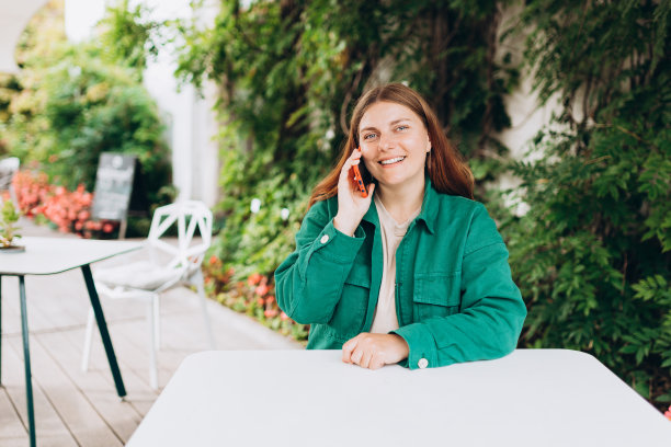 快乐的年轻女子等着点菜，坐在现代城市街道上的咖啡馆露台上。女孩讲着手机，温暖的晴天。图片下载