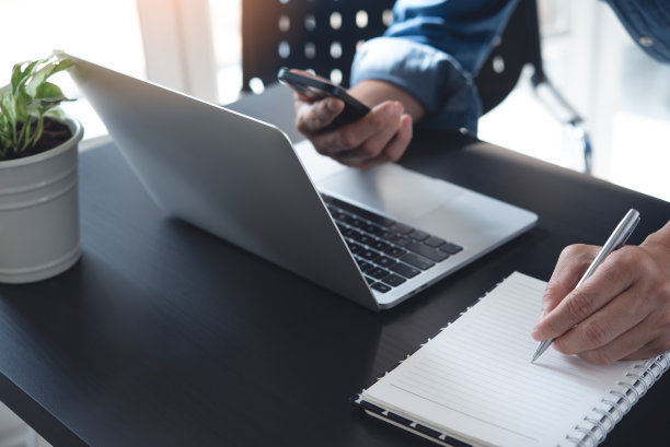 特写男人手拿笔在笔记本上写字，在笔记本电脑上工作，在办公桌上使用移动智能手机图片下载