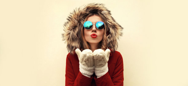 画像美丽的年轻女子吹她的嘴唇与红色的口红送甜蜜的空吻穿着夹克与毛皮帽在空白的背景图片下载