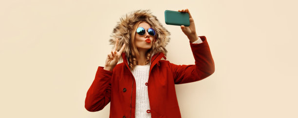 美丽的年轻女子的肖像，用智能手机自拍，吹她的嘴唇送吻，穿着红色夹克和皮草兜帽图片下载