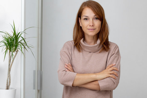 腰部向上美丽微笑的中年妇女双手交叉在胸前看着相机在浅棕色的衣服图片下载