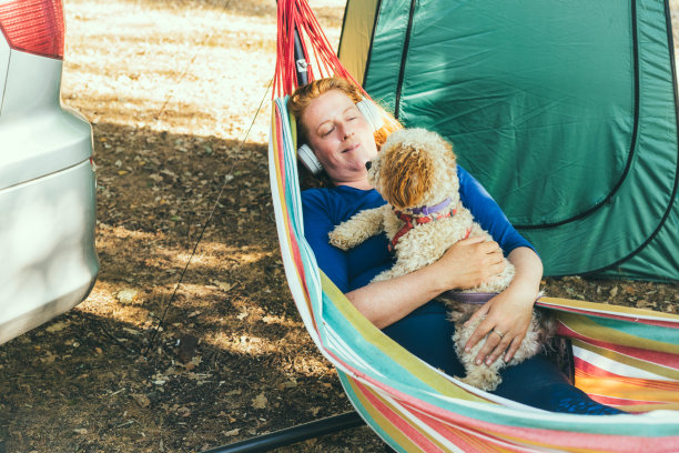 放松的女人听音乐与耳机躺在吊床与可卡普狗附近的房车露营之旅。女性和动物住在露营车上，周游世界。房车度假图片下载