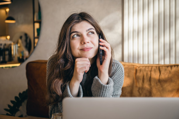 迷人的年轻女子一边打电话一边用笔记本电脑工作。图片下载