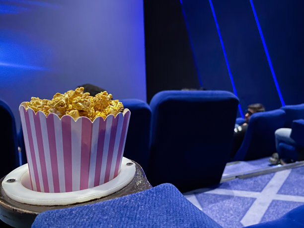 近距离图像的纸箱爆米花在电影院电影院座位杯托，电影零食食品浴缸，蓝色天鹅绒座椅，重点在前景图片下载