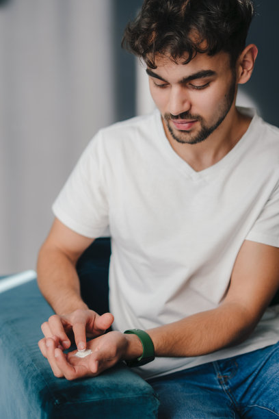 一个男人站在客厅的沙发上，手里拿着一滴要涂在脸上的面霜。美容产品。皮肤治疗。天然护肤化妆品。皮肤病、美容。身体保健。图片下载