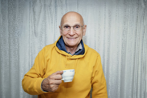 年长的退休男子戴着眼镜，拿着咖啡杯靠着白色的窗帘图片下载