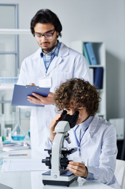 穿着实验服的年轻女人在显微镜下观察，她的助手在做笔记图片下载