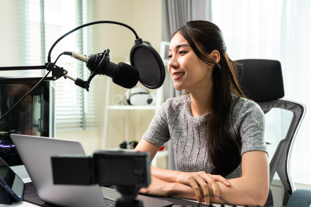 亚洲有吸引力的音频DJ女性对着麦克风说话广播。年轻美丽的女博主影响戴耳机和记录早上的新闻播客节目在家里的播音室。图片下载