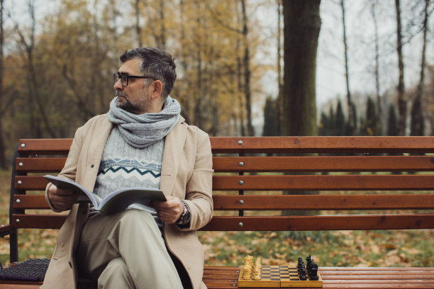 一位老人坐在秋天公园的长椅上看杂志。一个人在等他的朋友下棋。图片下载