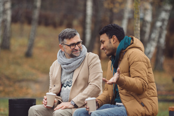 朋友，一个学长和一个年轻人坐在公园的长椅上，在秋天的公园里聊天。图片下载