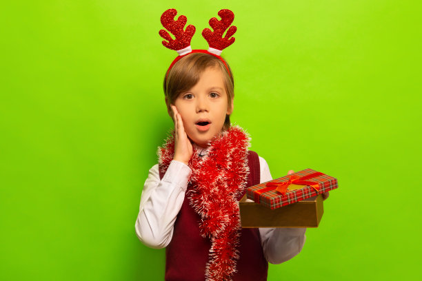 惊讶震惊的可爱男孩在红色圣诞驯鹿鹿角头饰与金属丝拿着打开的礼物，看着绿色背景上的相机。图片下载