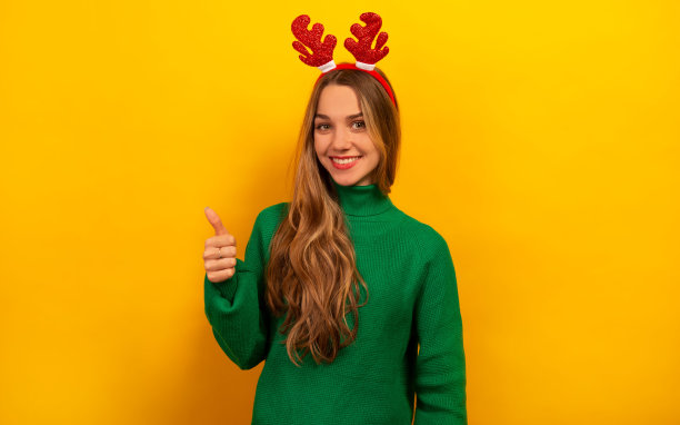 快乐微笑的女人竖起大拇指或一直竖起大拇指，看着黄色背景的相机。

快乐的褐发女孩，穿着绿色毛衣和红色圣诞驯鹿鹿角。图片下载