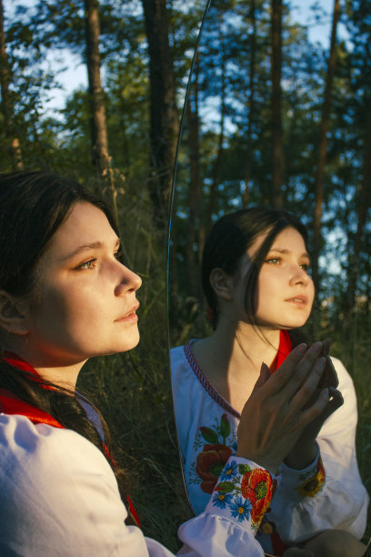 侧视年轻女子站在森林里镜子里的倒影附近，把她的手放在镜子表面图片下载