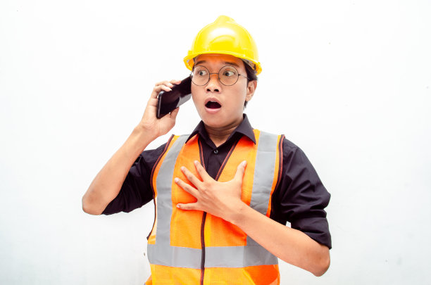 有吸引力的年轻亚洲男性建筑工人或工程师，橙色安全背心和黄色安全帽拿着电话，震惊，惊讶，哇的表情。图片下载