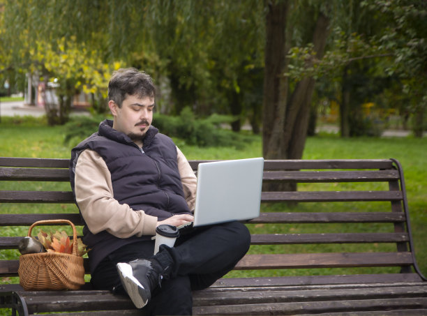 一个年轻人坐在公园的长椅上，手里拿着笔记本电脑。图片下载