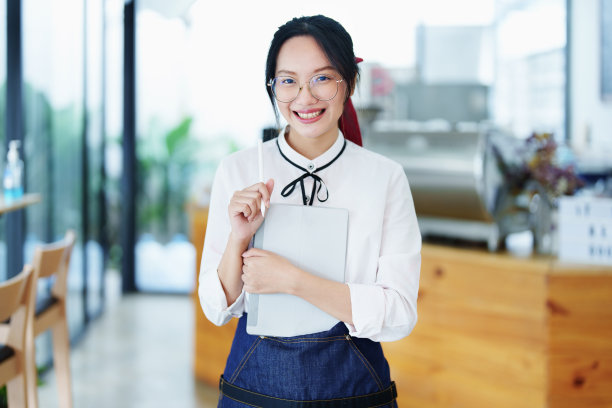 在咖啡店吧台前，一名年轻的亚洲女性用围裙系着平板电脑，脸上挂着微笑。企业主，餐厅，咖啡馆的概念图片下载