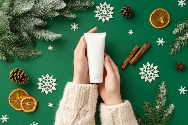 第一人称俯视图照片女性手在毛衣持有奶油管没有标签松枝球果干橙片肉桂棒雪花孤立的绿色背景与空白图片下载