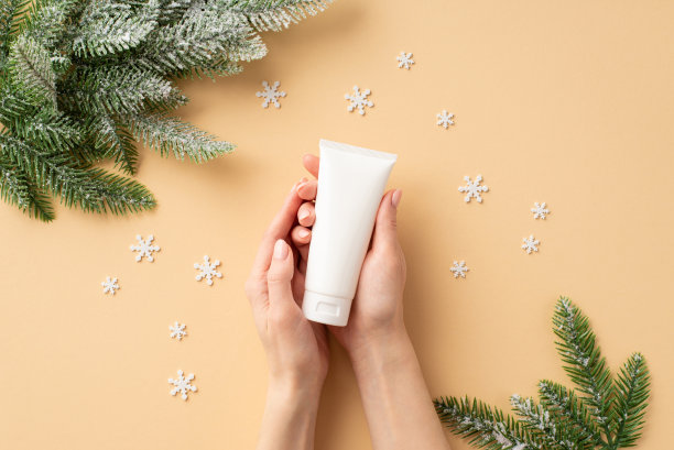 冬季护肤理念。第一人称俯视图照片的女性手拿白色管没有标签松枝在霜和雪花上孤立的淡色米色背景与copyspace图片下载