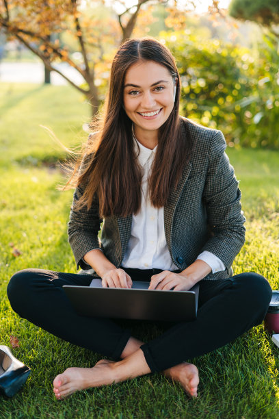 年轻的女商人学生自由职业者使用笔记本电脑为遥远的远程工作坐在绿草草坪上，看着相机和微笑。暑假。夏天的本性。绿色的大自然。互联网通信。图片下载