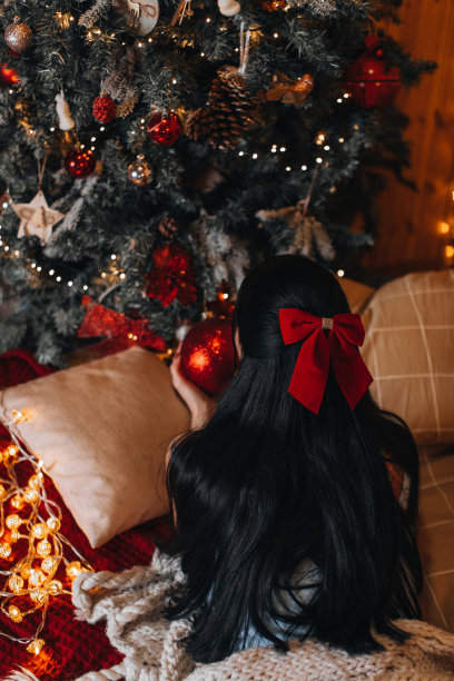 一位头发上扎着红蝴蝶结的褐发女子躺在挂满节日装饰的圣诞树旁。图片下载