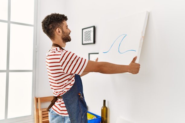 年轻的阿拉伯艺术家微笑着快乐地挂在艺术工作室的墙上画画布。图片下载