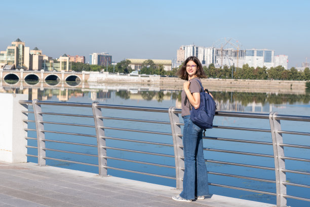 面带微笑的卷头发大学生背着背包站在城市的河堤上图片下载