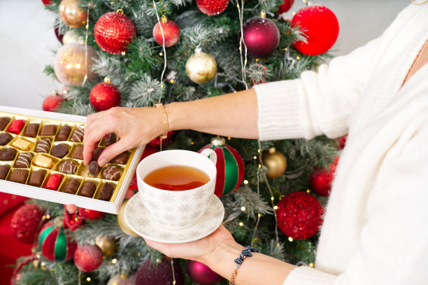 穿着白色毛衣的女孩在圣诞树的背景下端着一杯温暖的咖啡或茶。图片下载