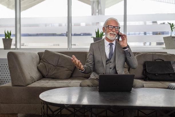 面带微笑的年长男性专业人士在办公室使用笔记本电脑时用智能手机讨论。图片下载
