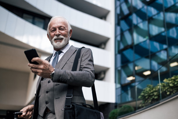 微笑的男性专业人士使用智能手机对抗现代建筑的画像。图片下载