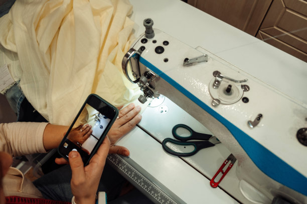 一名白人妇女在家庭作坊的缝纫机上缝制衣服，用智能手机用特写镜头录下了直播。小生意。博客的概念。图片下载