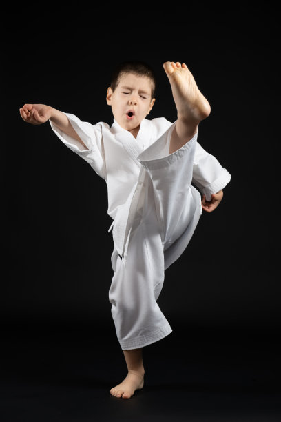 一个穿着和服的小男孩在黑色背景上练习空手道，踢腿向前。图片下载