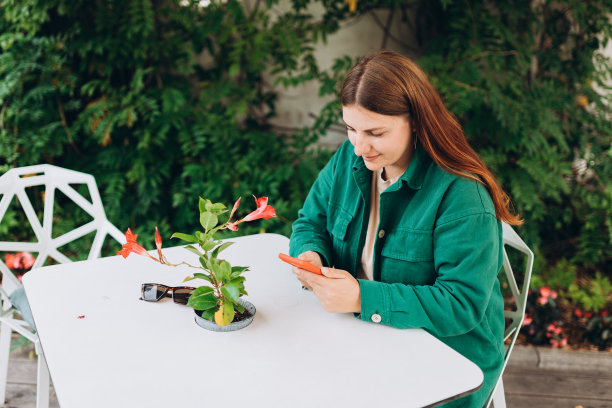 快乐的年轻女子与电话等待她的订单，坐在咖啡馆露台上的现代城市街道。电话沟通。城市生活方式的概念。旅行者，查看社交网络，发短信。图片下载