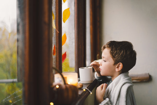 一个可爱的男孩裹着毯子喝着热茶，看着窗外美妙的秋色图片下载