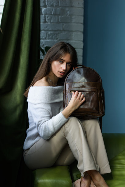 美丽苗条的女孩穿着米色的裤子和橄榄色的毛衣，坐在绿色的沙发上，背着一个闪闪发光的棕色小皮革背包图片下载