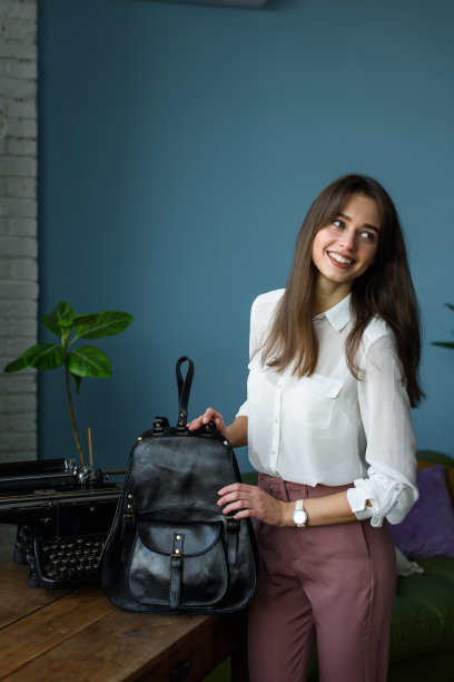 美丽苗条微笑的女孩在酒红色的裤子和一件白色衬衫摆姿势与黑色的皮革背包图片下载