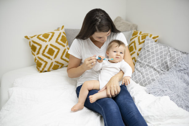 年轻漂亮的妈妈坐在床上打扫卫生，给宝宝按摩牙齿图片下载