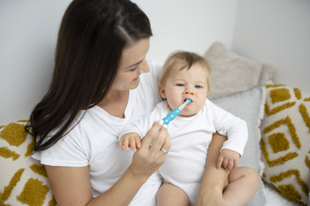 年轻漂亮的妈妈坐在床上打扫卫生，给宝宝按摩牙齿图片下载