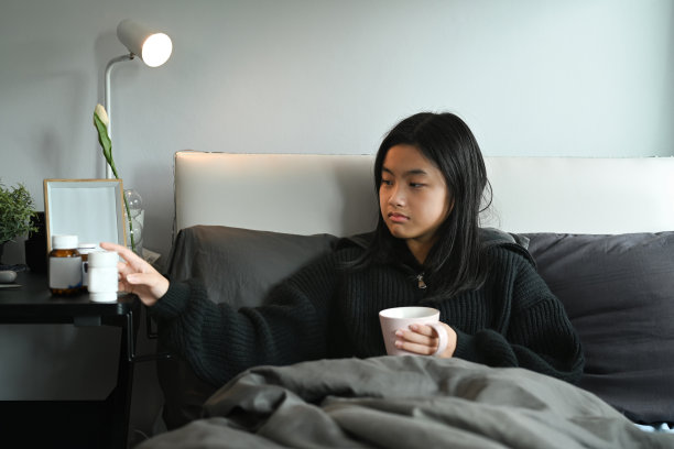 生病的年轻女子躺在床上，从床头柜里拿药来缓解症状。图片下载