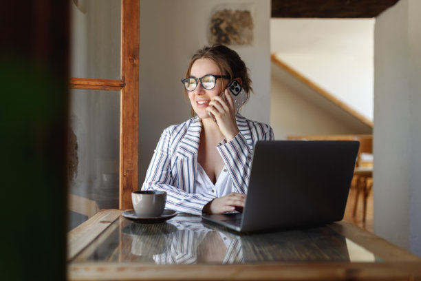 女人一边在办公室用笔记本电脑一边打电话，在窗户旁边喝咖啡图片下载