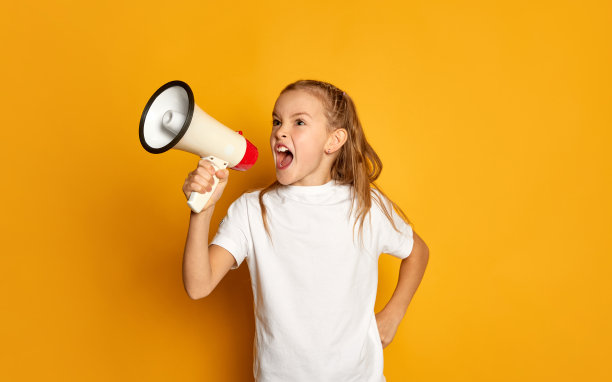 小美丽的女孩的肖像，孩子在白色的t恤摆姿势，叫喊在扩音器孤立在黄色的工作室背景图片下载